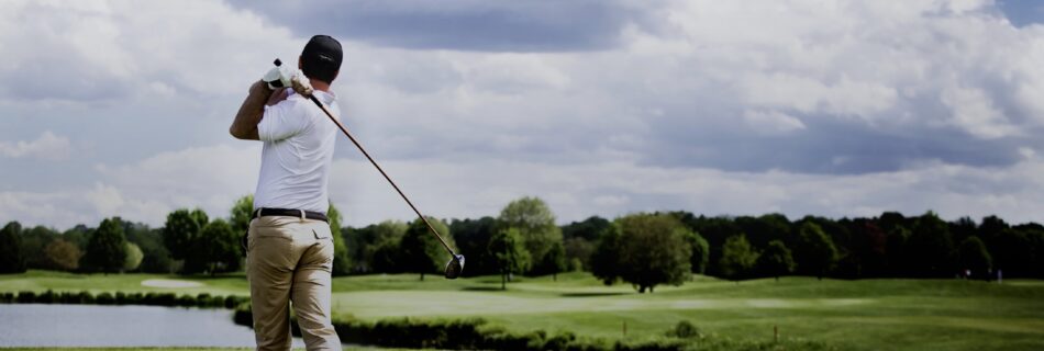 A golfer in mid-swing on a lush green golf course under a clear blue sky, with trees lining the fairway in the background. The golfer is wearing a white polo shirt, beige pants, and a white cap, focusing intensely on the shot. The pristine condition of the course, with neatly trimmed grass and a serene landscape, highlights the beauty of the golfing experience. The image captures the elegance and precision of the sport, emphasizing the golfer's form and the tranquil outdoor setting.