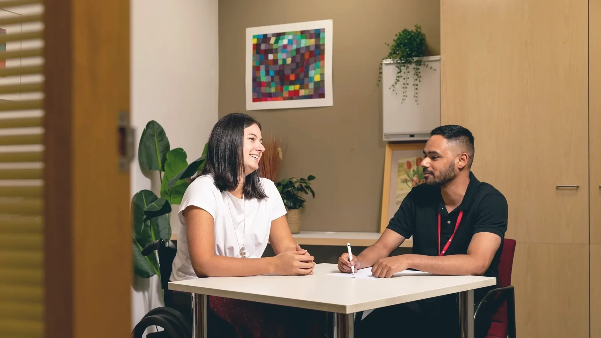 A man and a woman in a wheel chair engaged in conversation while seated at a table in a professional office setting.