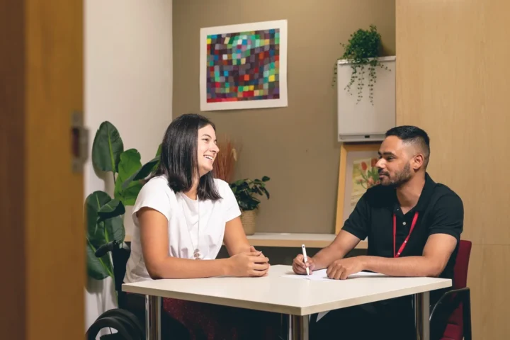 A man and a woman in a wheel chair engaged in conversation while seated at a table in a professional office setting.