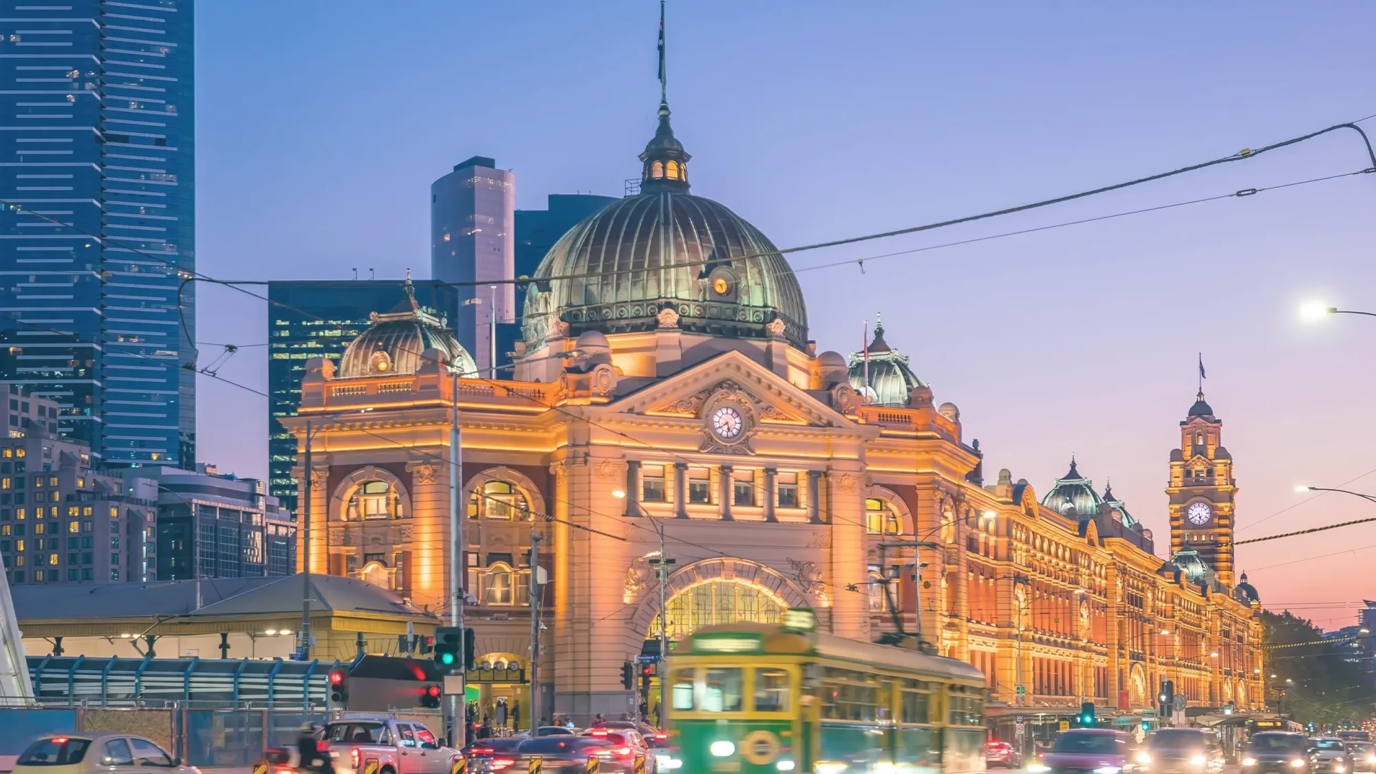 Bustling Melbourne city street at dusk, featuring traffic and a prominent building of Flinders Street Station illuminated by the evening light.