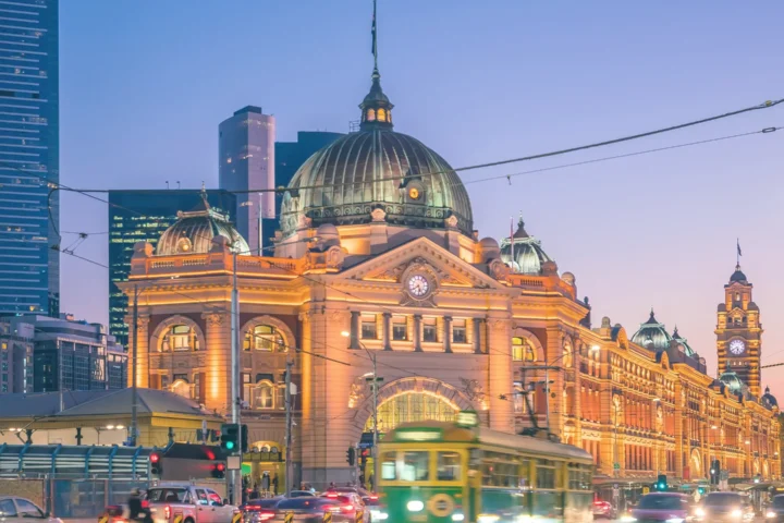 Bustling Melbourne city street at dusk, featuring traffic and a prominent building of Flinders Street Station illuminated by the evening light.