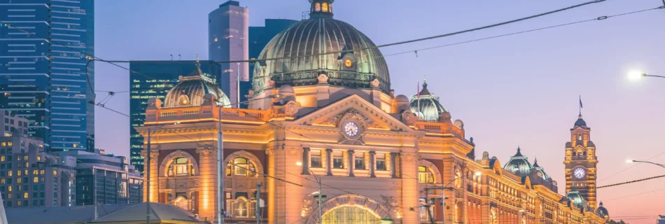 Bustling Melbourne city street at dusk, featuring traffic and a prominent building of Flinders Street Station illuminated by the evening light.