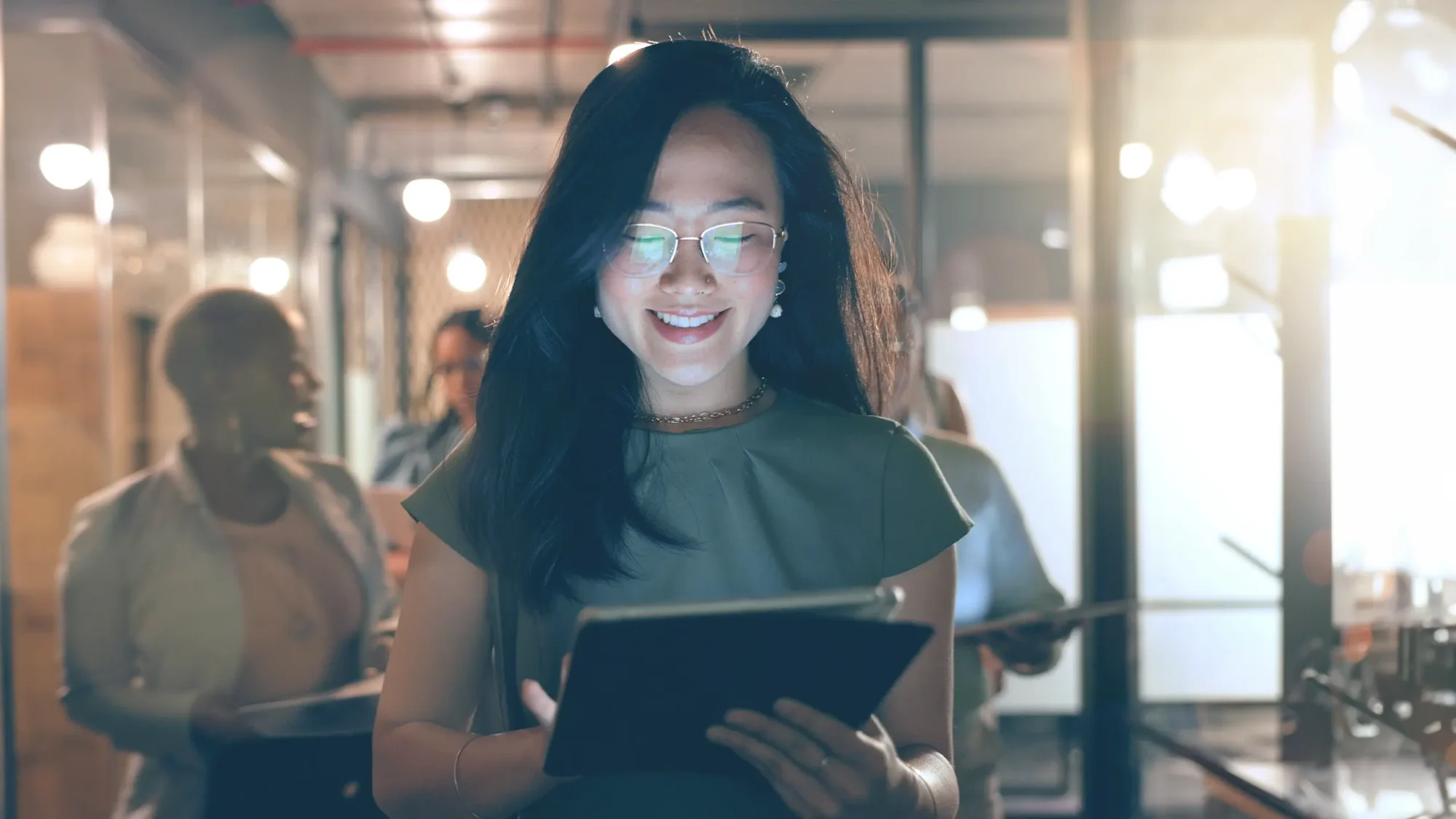 A woman wearing glasses is holding a tablet computer, focused on the screen with a thoughtful expression.