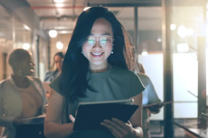 A woman wearing glasses is holding a tablet computer, focused on the screen with a thoughtful expression.