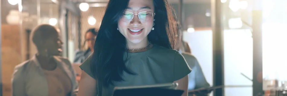 A woman wearing glasses is holding a tablet computer, focused on the screen with a thoughtful expression.