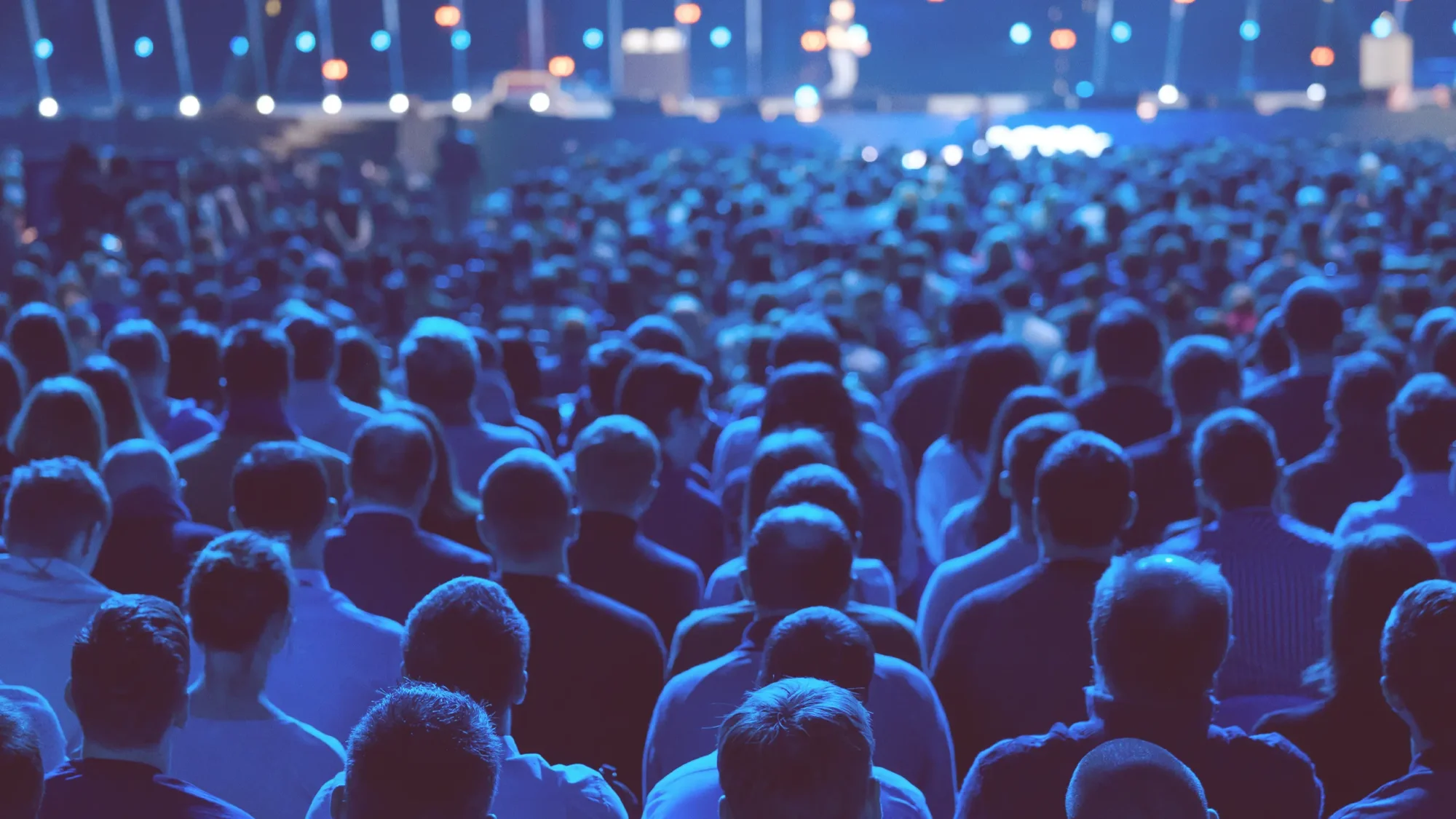 Audience from behind at a blue-lit event, watching a stage with soft focus lights.