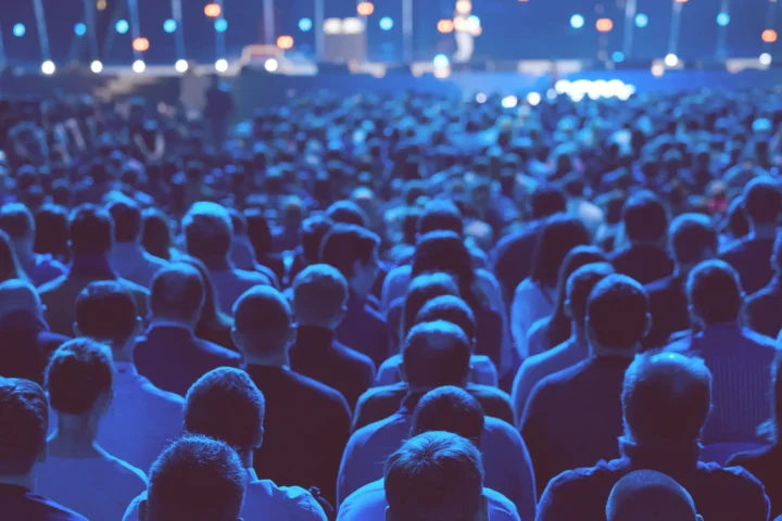 Audience from behind at a blue-lit event, watching a stage with soft focus lights.