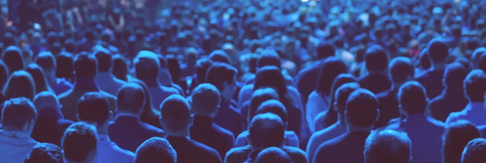 Audience from behind at a blue-lit event, watching a stage with soft focus lights.