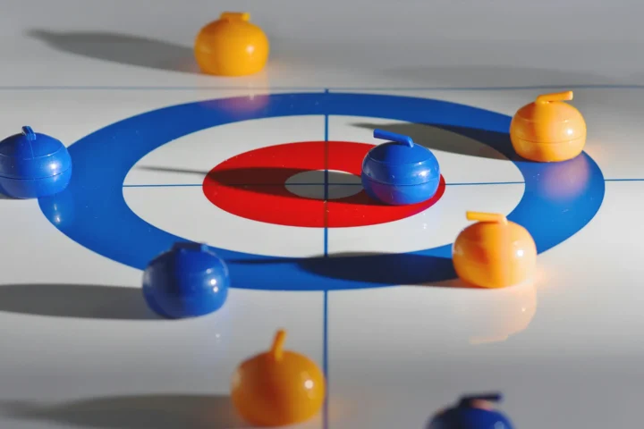 Mini curling game board with six stones, three orange and three blue, on a white surface with a red and blue target.