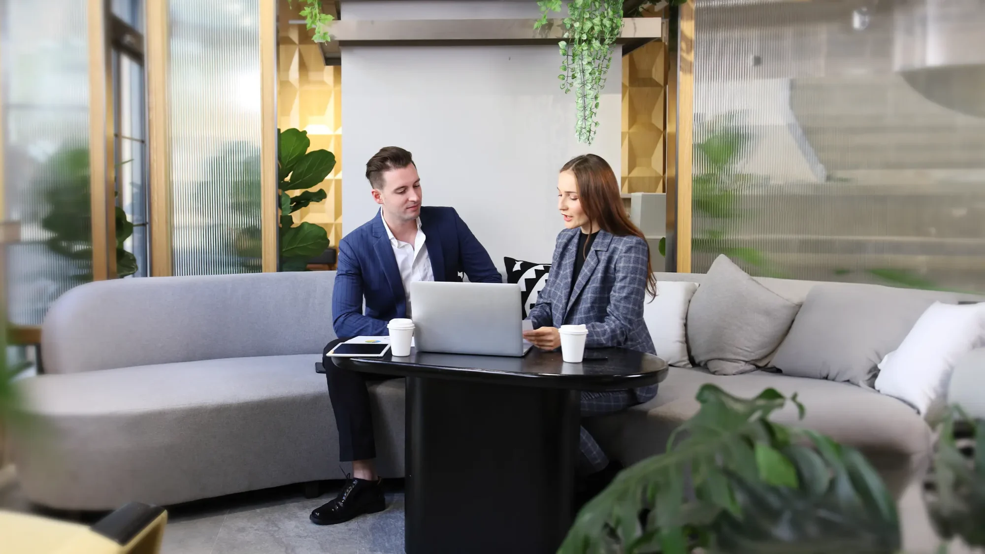 Two people in business attire sit on a sofa with a laptop and coffee cups on a table in a modern office setting talking about sustainability