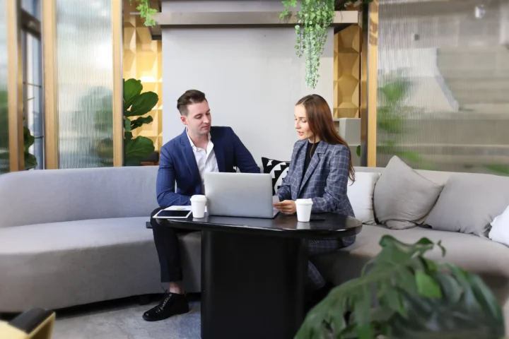 Two people in business attire sit on a sofa with a laptop and coffee cups on a table in a modern office setting talking about sustainability