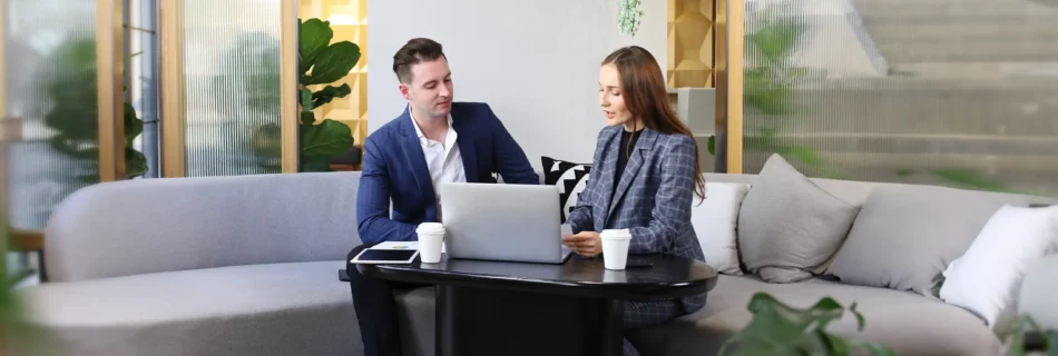 Two people in business attire sit on a sofa with a laptop and coffee cups on a table in a modern office setting talking about sustainability