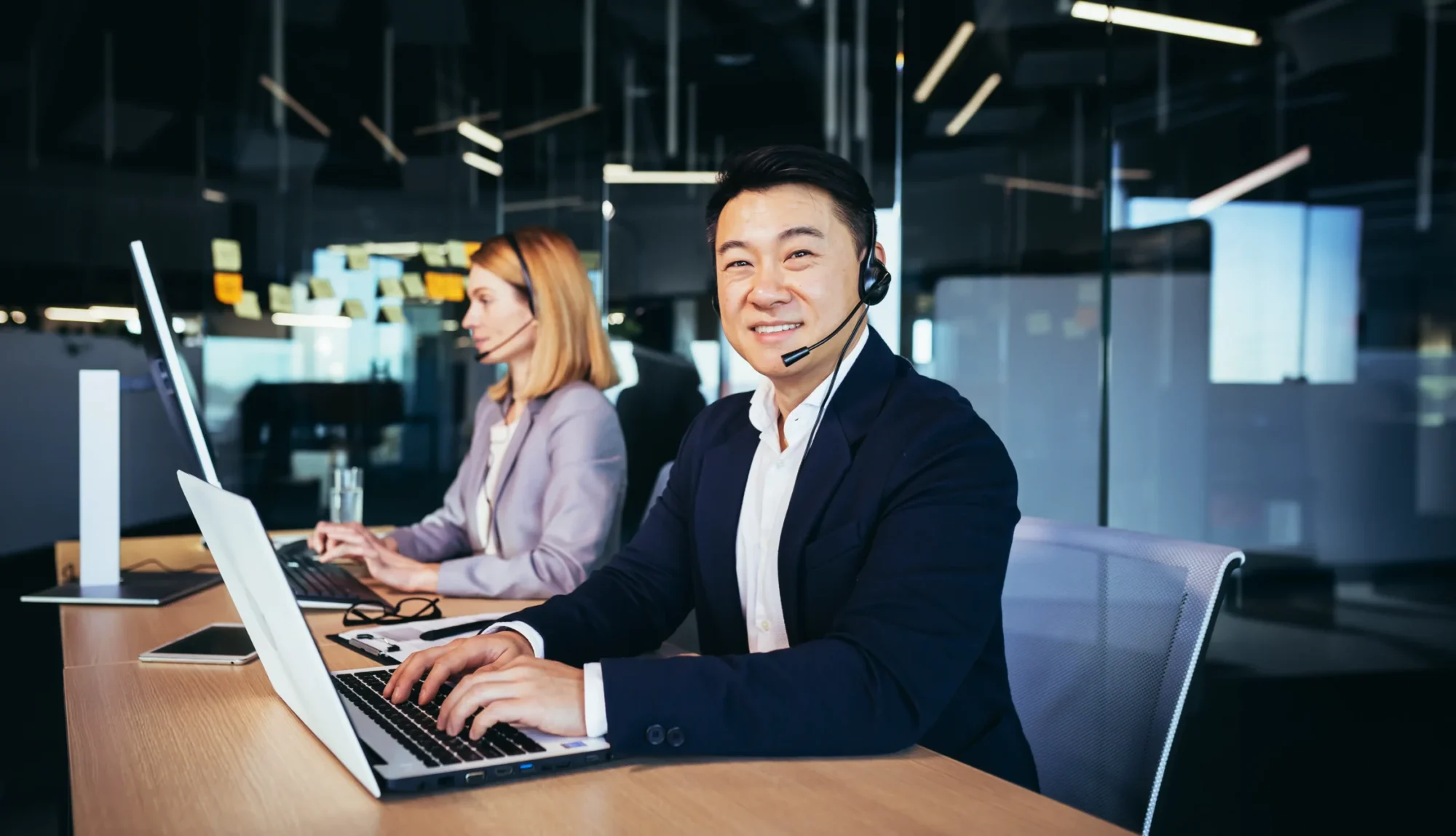 Axis Communications 2024 Contact centre with an asian man wearing a headset smiling and looking at the camera. A woman in the background wearing a headset working