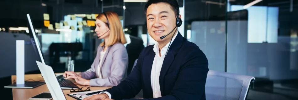 Axis Communications 2024 Contact centre with an asian man wearing a headset smiling and looking at the camera. A woman in the background wearing a headset working