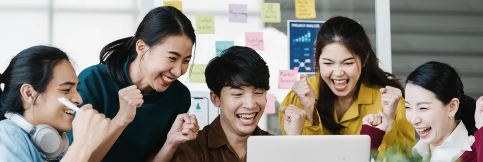 A group of five people employees gather around a laptop, smiling and celebrating. Behind them is a whiteboard with colorful sticky notes. Singapore