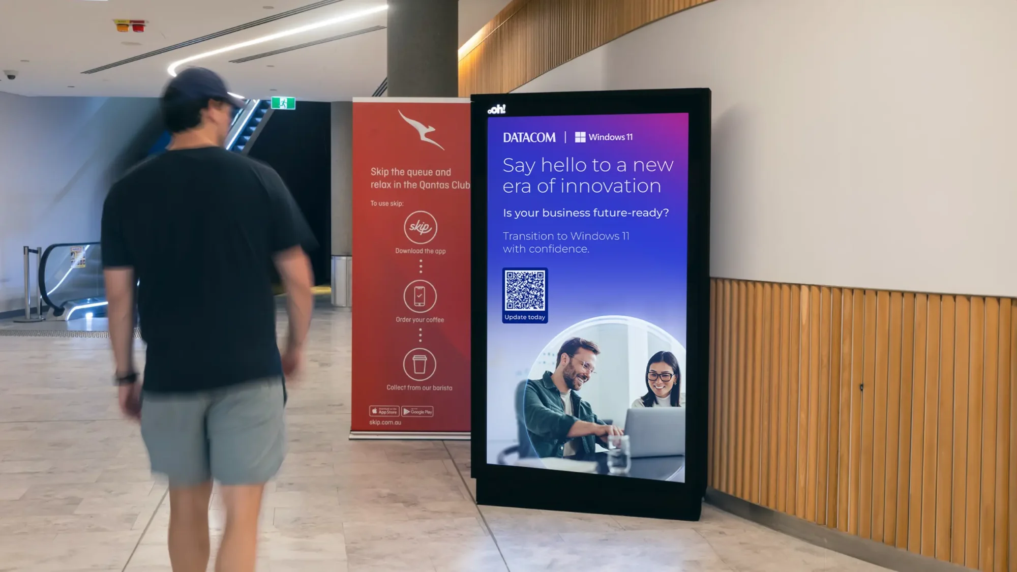 A digital advertising display in a modern indoor Qantas lounges shows a promotional message for Datacom and Windows 11. The ad features a headline reading "Say hello to a new era of innovation," with a subheading, "Is your business future-ready?" It encourages transitioning to Windows 11 with confidence and includes a QR code labeled "Update today." The ad features a photo of two smiling professionals working together on a laptop. Nearby, a smaller red sign promotes Qantas Club with steps to "Skip the queue," referencing an app for ordering coffee. An escalator and wooden wall panels are visible in the background.