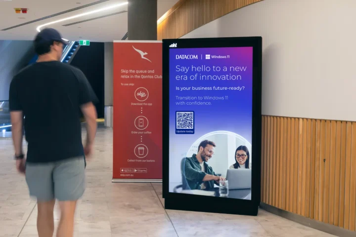 A digital advertising display in a modern indoor Qantas lounges shows a promotional message for Datacom and Windows 11. The ad features a headline reading "Say hello to a new era of innovation," with a subheading, "Is your business future-ready?" It encourages transitioning to Windows 11 with confidence and includes a QR code labeled "Update today." The ad features a photo of two smiling professionals working together on a laptop. Nearby, a smaller red sign promotes Qantas Club with steps to "Skip the queue," referencing an app for ordering coffee. An escalator and wooden wall panels are visible in the background.